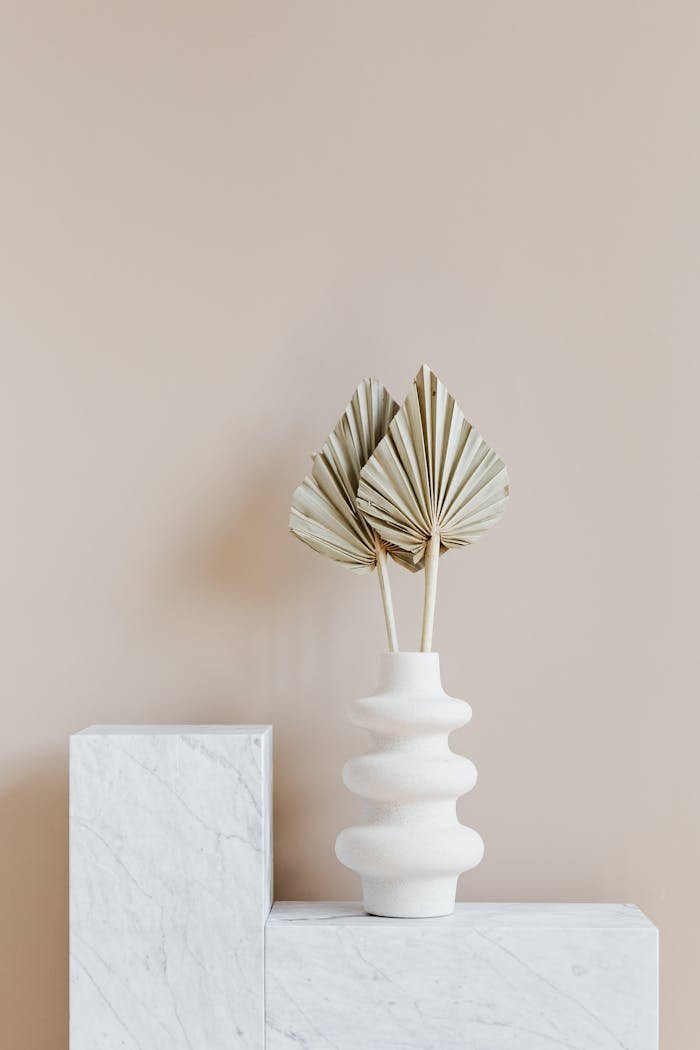 Decorative golden leaves in unique formed white vase placed on marble table against beige background