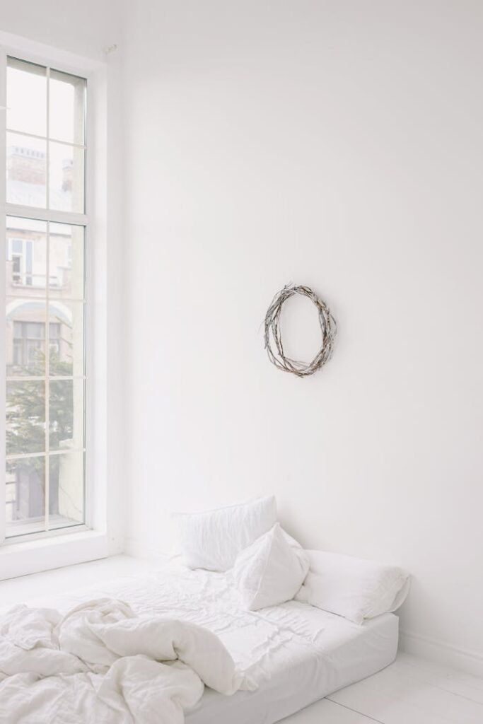Bright minimalist bedroom featuring a white bed, large window, and wall wreath.