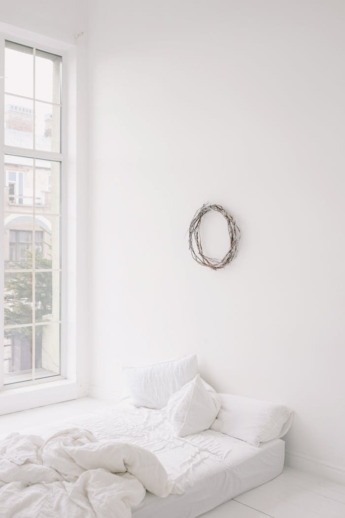 Bright minimalist bedroom featuring a white bed, large window, and wall wreath.
